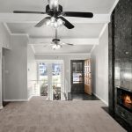 A spacious living room with beige carpet, a black marble fireplace, two ceiling fans, and large windows letting in natural light. An open kitchen is on the left and a wooden front door is visible in the background.