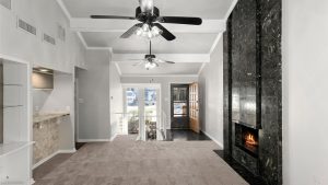 A spacious living room with beige carpet, a black marble fireplace, two ceiling fans, and large windows letting in natural light. An open kitchen is on the left and a wooden front door is visible in the background.