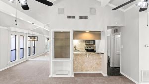 Modern open-concept living area with beige carpet, white walls, a built-in shelf, ceiling fans, and a small kitchen with stainless steel appliances and a tiled counter. Large windows provide ample natural light.