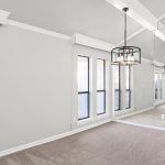 A bright, empty room with light gray walls, beige carpet, tall windows, a mirrored wall, and two modern black pendant lights hanging from a vaulted ceiling with white beams.