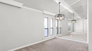A bright, empty room with light gray walls, beige carpet, tall windows, a mirrored wall, and two modern black pendant lights hanging from a vaulted ceiling with white beams.