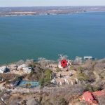 Aerial view of a lakeside neighborhood with a large body of water. Houses and trees line the shore. A red location pin marks one property near the lake. Tennis courts are visible to the left.