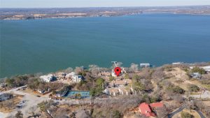 Aerial view of a lakeside neighborhood with a large body of water. Houses and trees line the shore. A red location pin marks one property near the lake. Tennis courts are visible to the left.