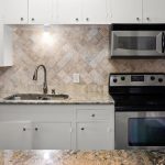 Modern kitchen with white cabinets, stainless steel appliances including a stove and microwave, a double sink with a gooseneck faucet, granite countertops, and a beige tile backsplash arranged in a diagonal pattern.