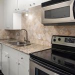 Modern kitchen with granite countertops, stainless steel oven and microwave, white cabinets, tiled backsplash, double sink, and bright lighting. To the left, large windows provide natural light and a view into a carpeted living area.