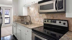 Modern kitchen with granite countertops, stainless steel oven and microwave, white cabinets, tiled backsplash, double sink, and bright lighting. To the left, large windows provide natural light and a view into a carpeted living area.