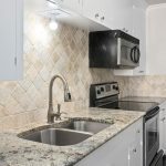 A modern kitchen with light cabinets, a granite countertop, stainless steel sink and appliances, tiled backsplash, and a view into a bathroom with a large mirror and double light fixture.