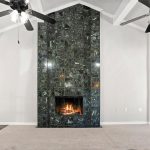 A modern living room with beige carpet, white walls, a polished dark marble fireplace with a lit fire, two black ceiling fans with lights, and a wooden door on the left.