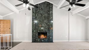 A modern living room with beige carpet, white walls, a polished dark marble fireplace with a lit fire, two black ceiling fans with lights, and a wooden door on the left.