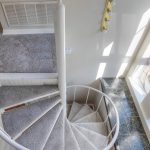 View from above of a carpeted spiral staircase with a white railing, next to large windows letting in sunlight, and a gray carpeted floor on the upper level.