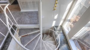 View from above of a carpeted spiral staircase with a white railing, next to large windows letting in sunlight, and a gray carpeted floor on the upper level.