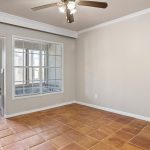 A small, empty room with terracotta tile flooring, beige walls, a ceiling fan with lights, a large window, and a doorway leading to another area with natural light.