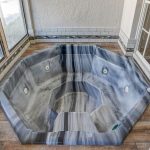 A gray marble-patterned octagonal jacuzzi tub is set in a tiled corner, next to large windows and glass doors, featuring built-in jets and surrounded by light-colored walls and floors.