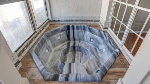 A gray marble-patterned octagonal jacuzzi tub is set in a tiled corner, next to large windows and glass doors, featuring built-in jets and surrounded by light-colored walls and floors.