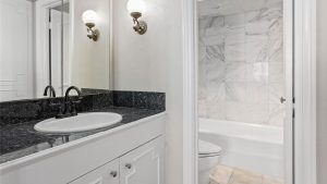 A bathroom with a white vanity, black granite countertop, and a sink with a black faucet. There’s a large mirror, wall-mounted light fixtures, and a bathtub with white and gray marble tile in the adjacent room.