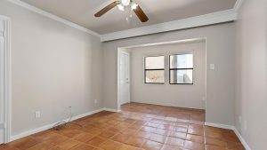 A bright, empty room with terracotta tile flooring, beige walls, white trim, two windows, a ceiling fan with lights, and a doorway leading to another area. Exposed wires are visible near one wall.