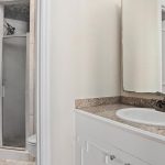 A bathroom with a small walk-in shower, a toilet, and a vanity with a brown granite countertop, white cabinets, an oval sink, and a mirrored medicine cabinet above. The walls are light-colored.