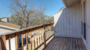 A small wooden balcony with a weathered floor and railing overlooks a residential area with leafless trees and neighboring houses under a clear blue sky.