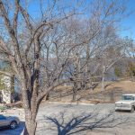 A paved lot with two parked vehicles, one white truck and one dark SUV, sits near leafless trees on a sunny day. In the background, there is a glimpse of a lake and hillside with sparse vegetation.