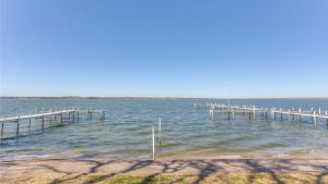 A calm lake under a clear blue sky with two wooden docks extending into the water from the shore. Shadows of tree branches are visible on the ground in the foreground.