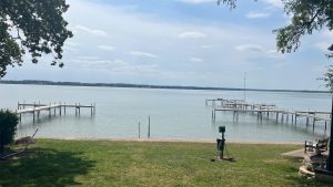 A peaceful lakeside scene with two wooden docks extending over calm water, grassy lawn in the foreground, and trees framing the view under a partly cloudy sky.