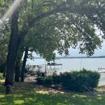 A sunbeam filters through tall trees onto a grassy lakeshore with a hammock, shrubs, and two wooden docks extending into the calm water under a partly cloudy sky.