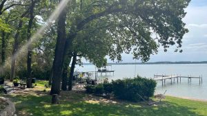 A sunbeam filters through tall trees onto a grassy lakeshore with a hammock, shrubs, and two wooden docks extending into the calm water under a partly cloudy sky.