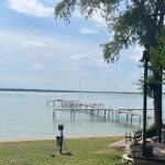 A calm lake with several wooden docks stretches into the water under a partly cloudy sky. In the foreground, there is a grassy lawn, patio furniture, and leafy trees on the right side.