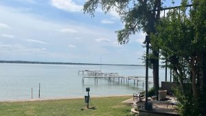 A calm lake with several wooden docks stretches into the water under a partly cloudy sky. In the foreground, there is a grassy lawn, patio furniture, and leafy trees on the right side.