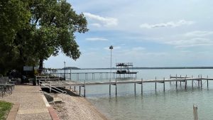 A lakeside scene with wooden docks extending over calm water, a tree providing shade on the left, patio chairs and tables along a paved walkway, and a two-level diving platform in the distance under a partly cloudy sky.