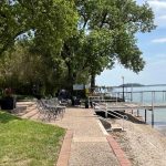 A paved pathway leads to a lakeside dock surrounded by trees, outdoor chairs, and tables. The calm lake is bordered by lush greenery and a partly cloudy sky is overhead.