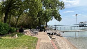 A paved pathway leads to a lakeside dock surrounded by trees, outdoor chairs, and tables. The calm lake is bordered by lush greenery and a partly cloudy sky is overhead.