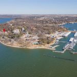 Aerial view of a peninsula with a marina filled with boats, waterfront homes, docks, and trees. A red location marker highlights one of the houses near the shoreline on the left side of the image.