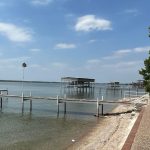 A lakeside scene with wooden docks extending into the calm water, a sandy shore with a brick walkway, a house on the right, and blue sky with scattered clouds overhead.
