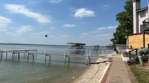 A lakeside scene with wooden docks extending into the calm water, a sandy shore with a brick walkway, a house on the right, and blue sky with scattered clouds overhead.