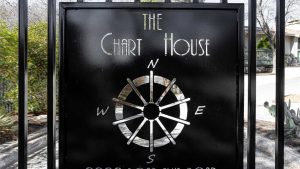 A black metal sign for "The Chart House" features a nautical compass design with N, S, E, and W directions and is attached to a gate with plants and a building visible in the background.