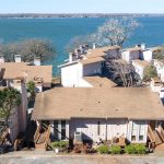 Aerial view of beige lakeside townhouses with brown roofs, surrounded by bare trees, situated along the edge of a large blue lake under a clear sky.