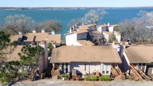 Aerial view of beige lakeside townhouses with brown roofs, surrounded by bare trees, situated along the edge of a large blue lake under a clear sky.