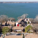 Aerial view of a lakeside residential area with multiple houses and trees. A red location pin marks one of the houses near the shoreline, with a large body of water and docks visible in the background.