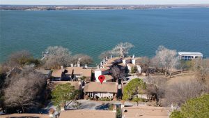 Aerial view of a lakeside residential area with multiple houses and trees. A red location pin marks one of the houses near the shoreline, with a large body of water and docks visible in the background.
