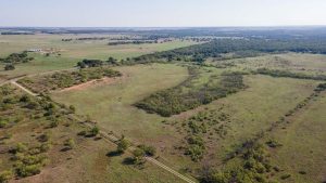 Aerial view of a vast rural landscape with grassy fields, scattered trees, dirt roads, and distant hills under a clear sky. A few small buildings are visible in the far distance.