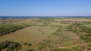 A wide aerial view of rolling, grassy fields with scattered trees under a clear blue sky, stretching to the horizon. No buildings are visible; the landscape appears rural and open.