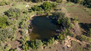 A small, dark pond surrounded by green trees and shrubs in a grassy, slightly hilly landscape, seen from above on a sunny day.