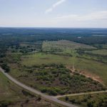 Aerial view of a rural landscape with open fields, patches of trees, and a winding road running through grassy land under a clear sky.