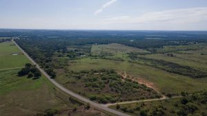 Aerial view of a rural landscape with open fields, patches of trees, and a winding road running through grassy land under a clear sky.