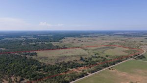 Aerial view of a large, open rural landscape with fields, trees, and scattered vegetation. A red outline marks the boundary of a specific parcel of land. A road runs along one side of the outlined property.