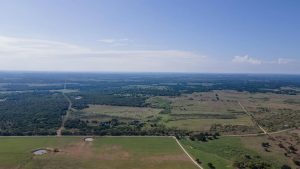 Aerial view of a vast rural landscape with green fields, patches of trees, small ponds, dirt roads, and a clear blue sky stretching across the horizon.
