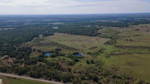 Aerial view of a vast rural landscape with open green fields, scattered trees, small ponds, and a road running along the bottom edge under a partly cloudy sky.