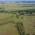 Aerial view of a rural, grassy landscape with patches of trees, a small pond, scattered houses, and farmland stretching into the distance under a clear sky.