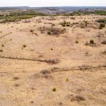 Aerial view of the historic S&S Ranch reveals a dry, grassy landscape with sparse shrubs and a partially collapsed stone or concrete fence forming an L-shape, surrounded by open fields and rolling hills in the distance.
